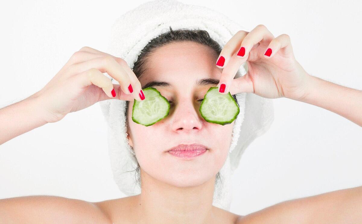 Young woman applying cucumber slices over her eyes against white background