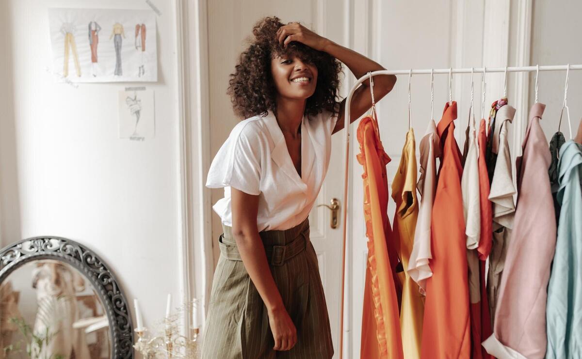 Curly young darkskinned woman in stylish white summer blouse and khaki shorts smiles sincerely and leans on hanger in white room