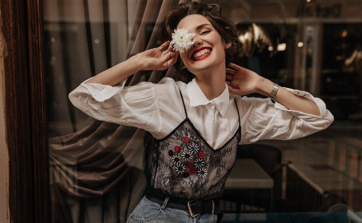 Positive woman with short hair in long sleeve white and black shirt smiling sincerely in cafe. Cheerful lady in jeans holding flower inside.