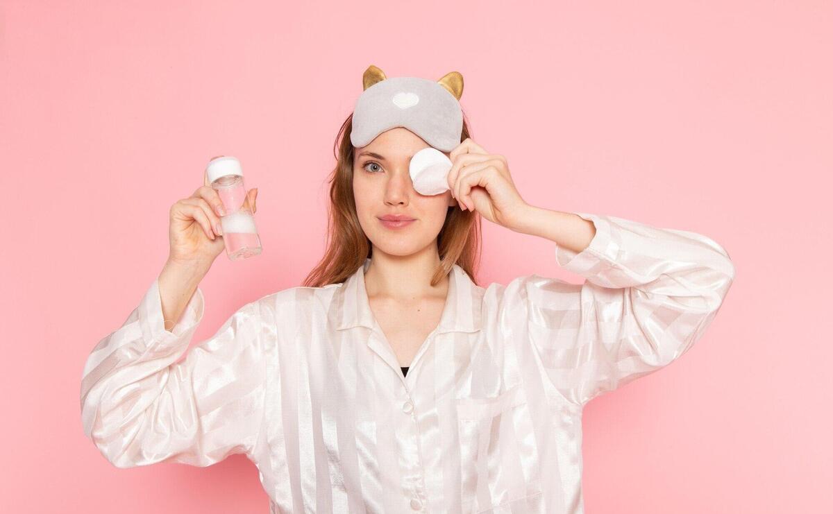 young female in pajamas and sleep mask cleaning her face up with spray on pink