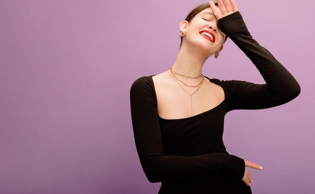 Pretty young caucasian brunette woman in black blouse with closed eyes keeps hand on face smiling on purple background