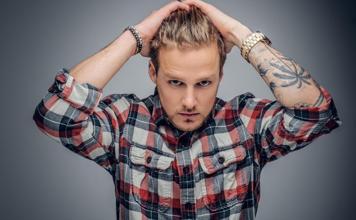 Portrait of a blond male with tattooed arms on his head dressed in a plaid shirt isolated on grey vignette background.