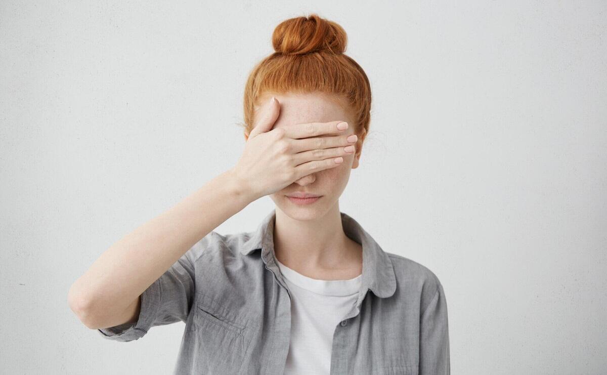 redhead girl with hair bun hiding eyes under hand while feeling ashamed. Serious young woman in causal clothing covering face with hand. Human facial expressions and emotions