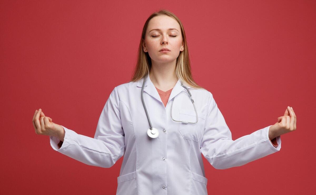 Peaceful young blonde female doctor wearing medical robe and stethoscope around neck meditating with closed eyes 