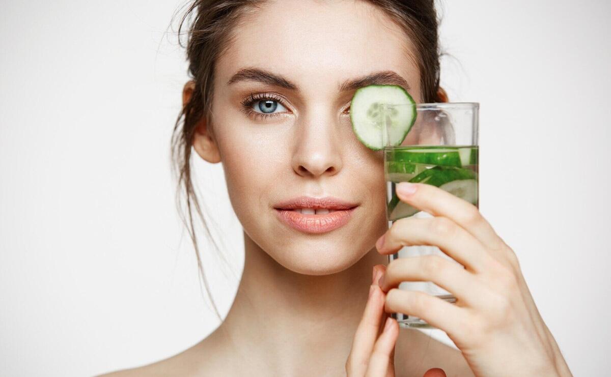 Close up of beautiful nude girl smiling looking at camera holding glass of water with cucumber slices over white background. Healthy nutrition. Beauty and skincare.