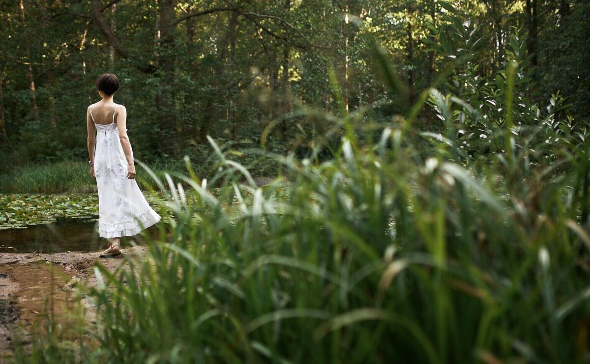 Outdoor summertime image of romantic adorable young female wearing long white dress relaxing in wild nature alone on weekend, standing by pond in background with fresh green grass in foreground