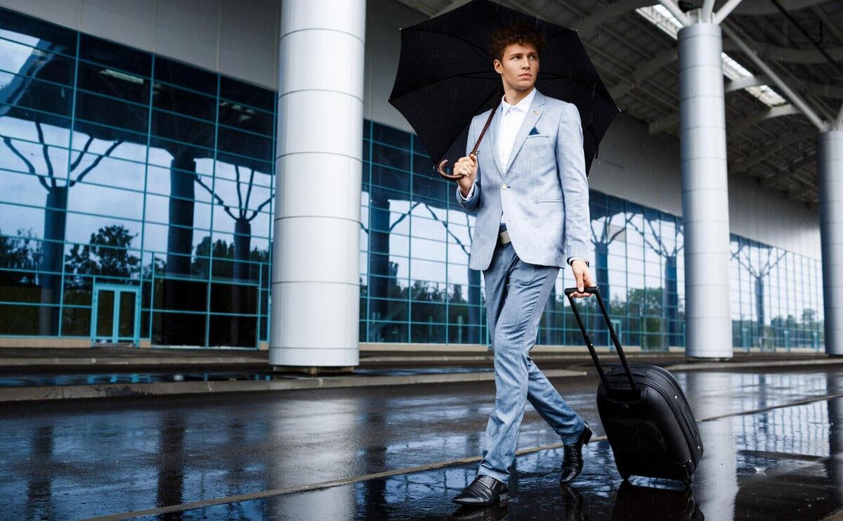 Picture of  young redhaired businessman holding black umbrella and suitcase walking in rain at airport