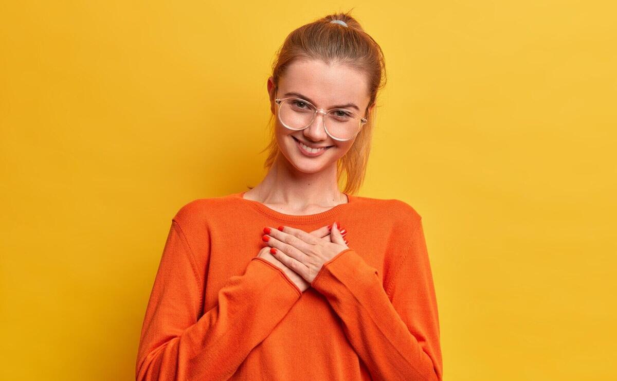 Cute pleased satisfied girl presses hands to heart, appreciates nice gift, looks gratefully being full of love and kindness smiles tenderly wears optical glasses, orange jumper stands indoor