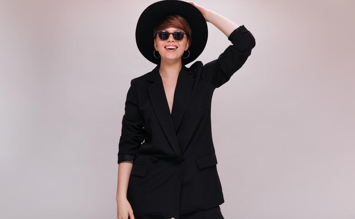 Portrait of stylish lady in sunglasses and wide brimmed hat. Cool young woman in black jacket and pants poses and smiles on isolated background