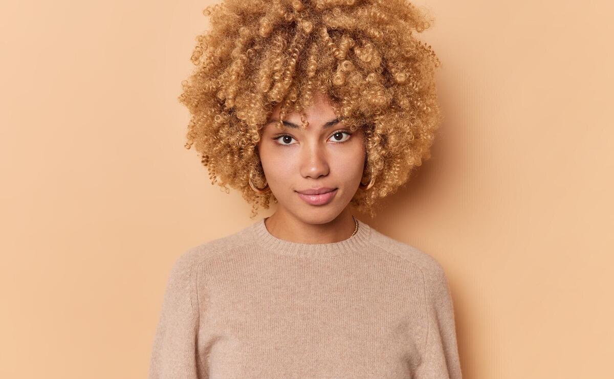 Portrait of serious confident sassy good looking woman with bushy curly hair looks directly at camera stands indoor against beige background wears casual jumper. Human face expressions concept