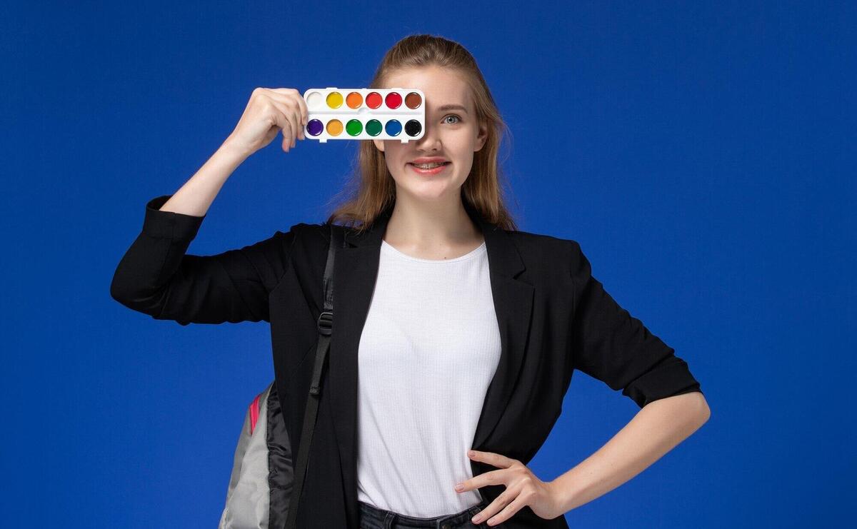 Front view female student in black jacket wearing backpack holding paints for drawing on blue wall drawing art college university