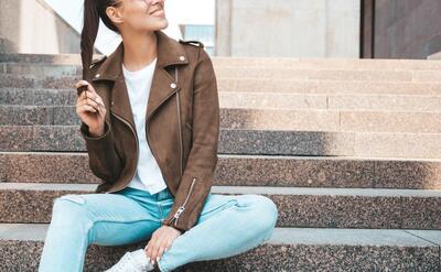 Portrait of beautiful smiling brunette model dressed in summer hipster jacket and jeans clothes. Trendy girl sitting on steps in the street background. Funny and positive woman in round sunglasses