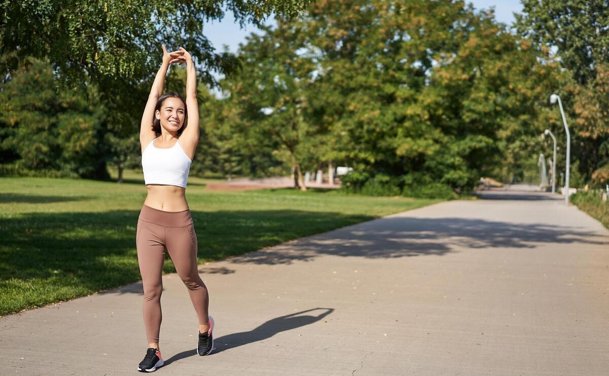 Smiling asian girl stretching after good workout in park listening music in wireless headphones